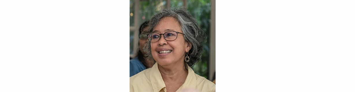 Portrait of Suriani Suratman, woman in a yellow shirt with greying shoulder length hair, smiling